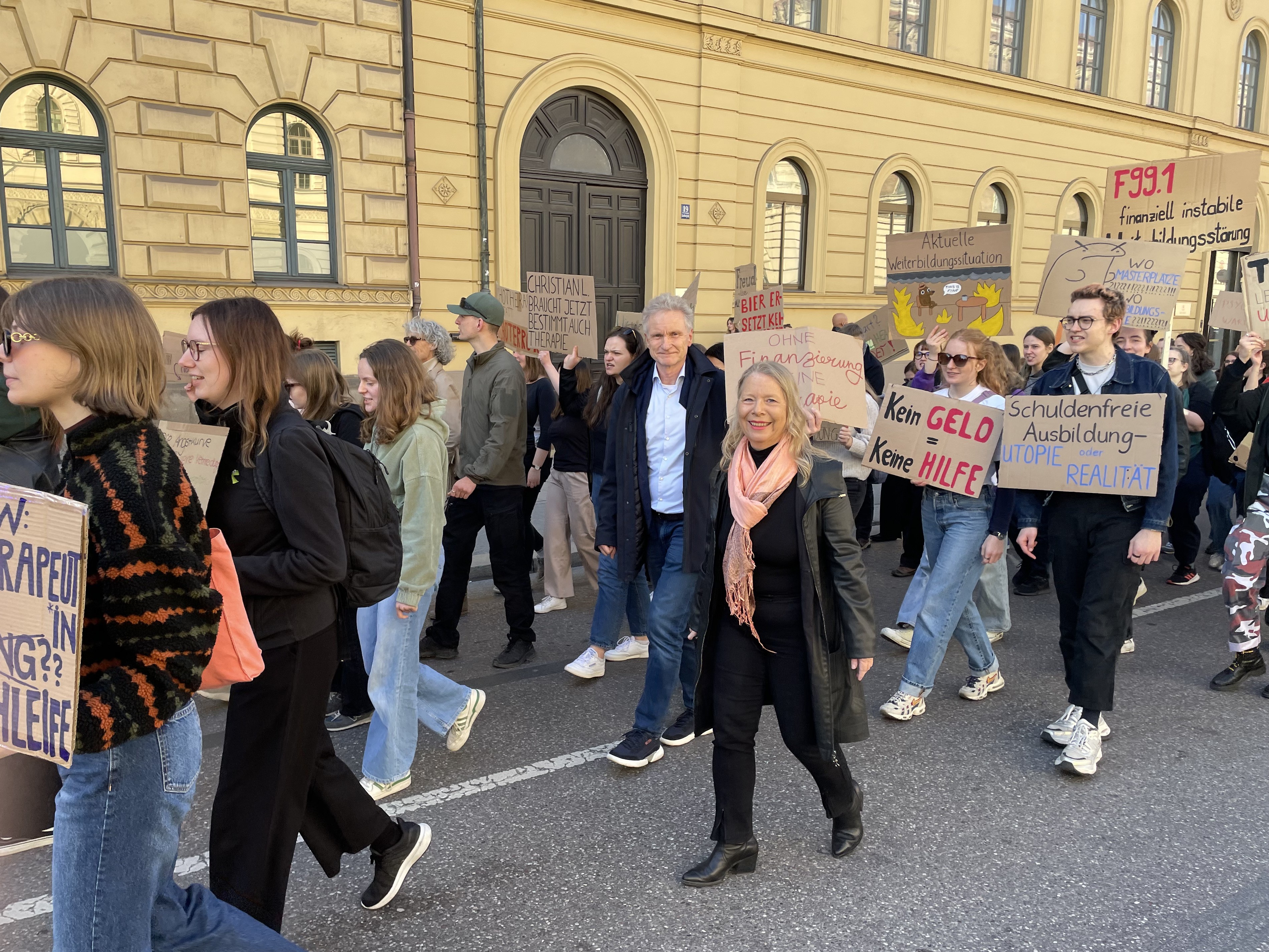 Demonstierende mit selbstgemalten Plakaten laufen bei einer Demonstration mit. Mitten drin sind Herr Melcop und Frau Pielsticker.